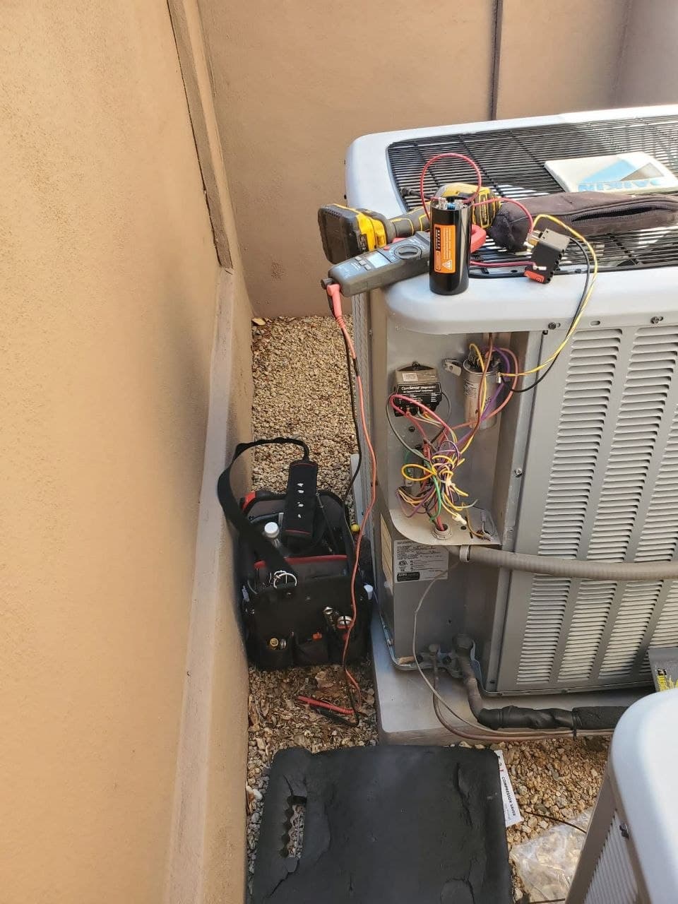Meter leads clipped to the contactor inside an open Daikin condenser, with a technician's tool bag on the ground beside it